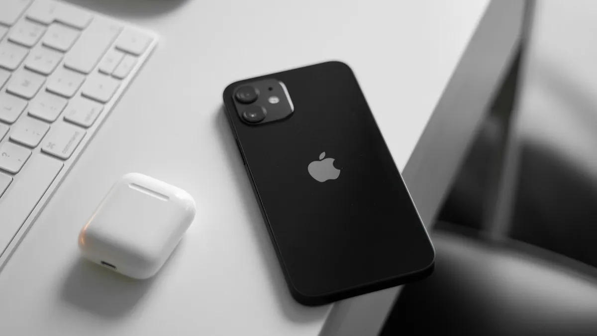 A black iPhone on a white desk next to a white AirPods case